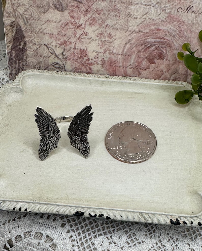 Op de afbeelding: Een zilveren ring met twee engelenvleugels aan weerszijden. De ring ligt op een wit dienblad met een Amerikaanse kwart voor de schaal.