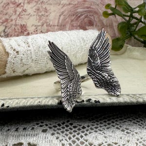 Op de afbeelding: Een ring met twee zilveren engelenvleugels. De vleugels zijn gedetailleerd en hebben een textuur.