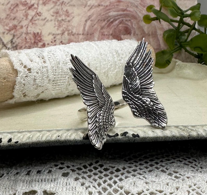 Op de afbeelding: Een ring met twee zilveren engelenvleugels. De vleugels zijn gedetailleerd en hebben een textuur.
