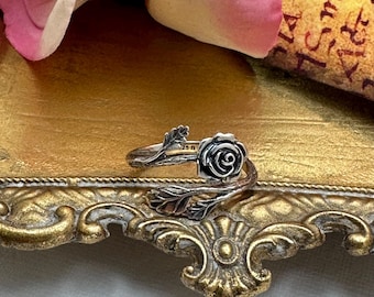 May include: A silver ring with a rose design. The ring wraps around the finger with a detailed rose blossom and leaf accents. The ring is displayed on a gold surface with an ornate border.