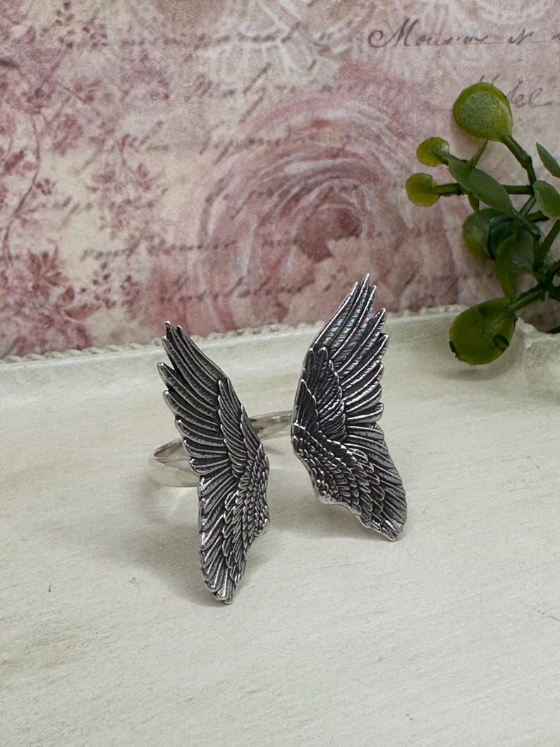 Op de afbeelding: Een paar zilveren ringen met engelenvleugels. De vleugels zijn gedetailleerd en hebben een textuur.