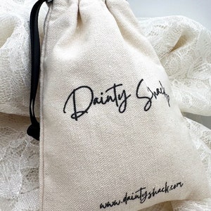 Op de afbeelding: Een witte katoenen trekkoordtas met zwarte trekkoorden. De tas heeft zwarte tekst die "Dainty Snack" en "www.daintyshack.com" zegt.