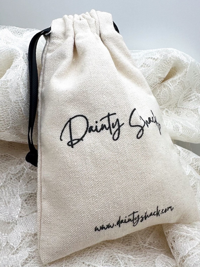 Op de afbeelding: Een witte katoenen trekkoordtas met zwarte trekkoorden. De tas heeft zwarte tekst die "Dainty Snack" en "www.daintyshack.com" zegt.