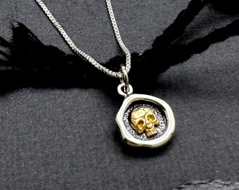 Op de afbeelding: Een zilveren ketting met een zilveren hanger met een gouden schedelontwerp. De hanger is gevormd als een waszegel.