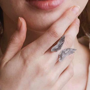 Op de afbeelding: Een zilveren ring met twee vleugels. De vleugels zijn gedetailleerd en hebben een textuur.