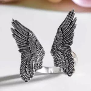 Op de afbeelding: Een zilveren ring met aan weerszijden twee engelenvleugels. De vleugels zijn gedetailleerd en hebben een textuur.