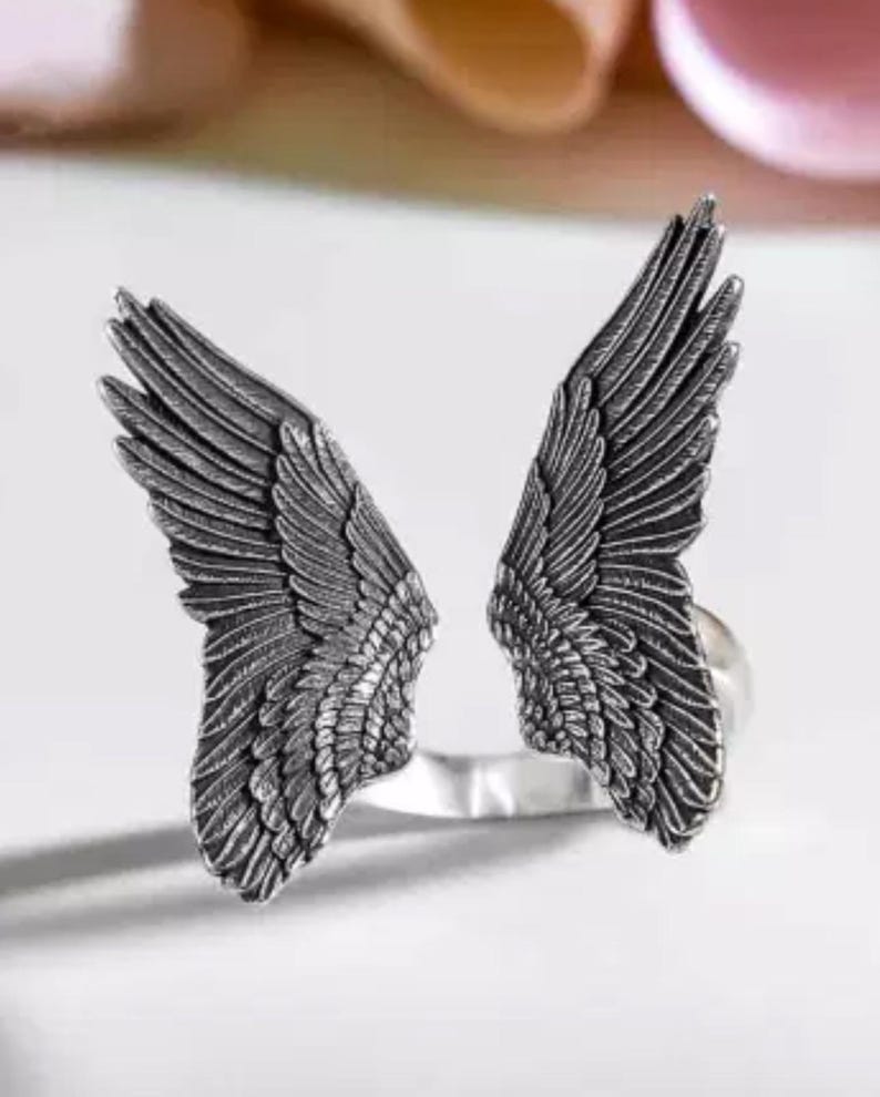 Op de afbeelding: Een zilveren ring met aan weerszijden twee engelenvleugels. De vleugels zijn gedetailleerd en hebben een textuur.