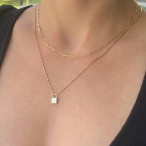 May include: Two gold necklaces layered on a person's neck. The top necklace is a chain with oval links. The bottom necklace has a small padlock pendant.