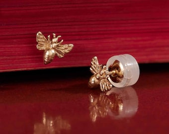 May include: A pair of gold bee stud earrings. The earrings are small and delicate, with a detailed bee design. The earrings are on a red surface.
