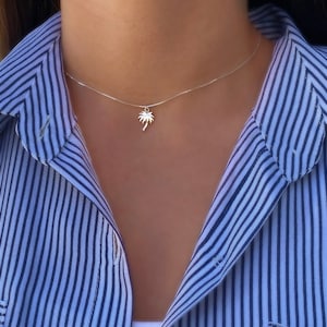 May include: A silver necklace with a palm tree pendant. The necklace is on a person wearing a blue and white striped shirt.