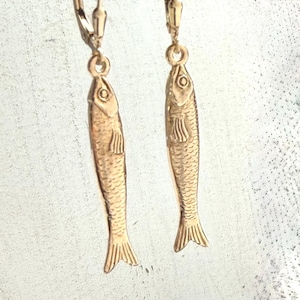 May include: A pair of gold-colored fish-shaped earrings. Each earring features a detailed fish design with scales, fins, and a tail. The earrings hang from lever-back closures.