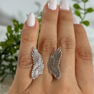 Op de afbeelding: Een zilveren ring met een gedetailleerd vleugelontwerp. De ring wordt gedragen aan de wijsvinger van een hand met witte nagellak.