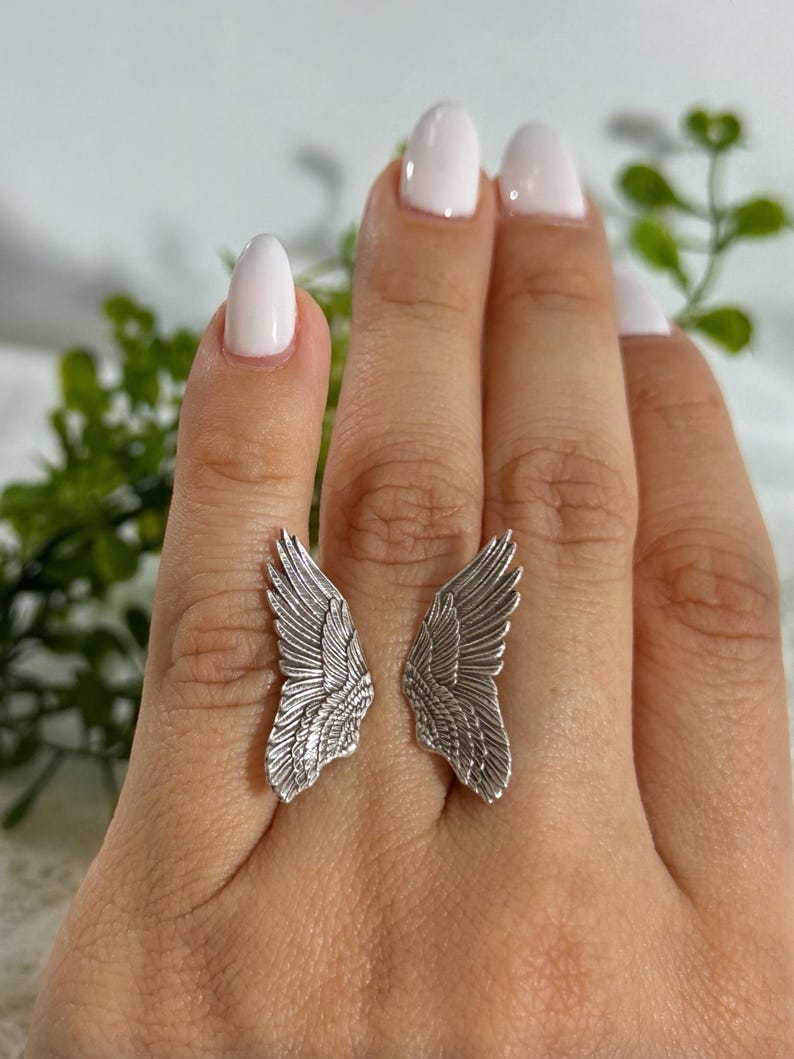 Op de afbeelding: Een zilveren ring met een gedetailleerd vleugelontwerp. De ring wordt gedragen aan de wijsvinger van een hand met witte nagellak.