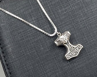 May include: A silver chain necklace with a silver Mjolnir pendant. The pendant is detailed with intricate carvings and a raised hammer head.