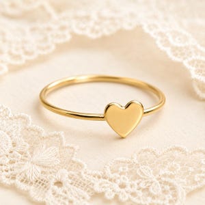 May include: A thin gold ring featuring a small, smooth, solid heart-shaped charm centered on the band. The ring has a polished, reflective metallic finish and a simple, delicate construction with a rounded band profile and a flat heart design.
