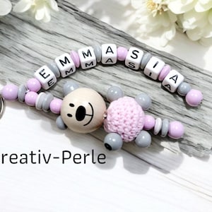 Könnte beinhalten: Ein rosa-grauer Perlen-Schlüsselanhänger mit einem Holz-Bären-Anhänger und dem Namen "EMMASIA" in weißen Buchstaben.