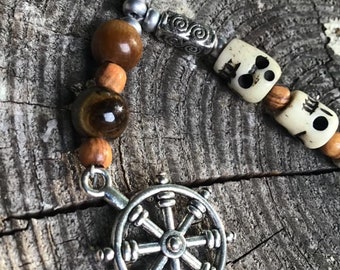May include: A silver charm necklace with a wooden bead and a silver steering wheel charm. The necklace also features a tiger's eye bead and a skull bead.