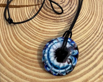 May include: A blue and white glass pendant with black swirls, hanging from a black leather cord. The pendant is round and has a white center with blue and black swirls around it.