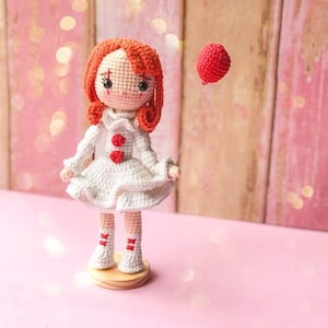 May include: A handmade crochet doll with red hair and a white dress, standing on a wooden base. The doll has a red balloon floating nearby. The doll's features include black eyes, rosy cheeks, and red accents on the dress and socks. The background is a soft pink.