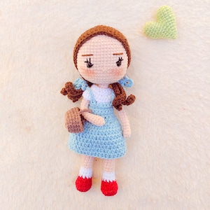 May include: A crocheted doll with brown hair, a blue dress, and red shoes. The doll is holding a brown crocheted purse. A green crocheted heart is in the background.