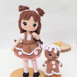 May include: Two handmade crochet dolls. One is a girl doll with brown hair, a brown dress, and pink accents. The other is a gingerbread man doll with white frosting details. Both dolls are standing on wooden bases.