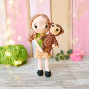 以下が含まれることがあります： 茶色の髪と緑のシャツを着た編みぐるみの女の子が、編みぐるみの猿を抱っこしています。女の子は茶色のショートパンツと黒い靴を履いています。女の子と猿は木製の表面に立っています。