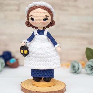 May include: A crocheted doll with brown hair, wearing a white cap, a navy blue long-sleeved dress, and a white apron. The doll has large black eyes and holds a small black lantern with a yellow light. It stands on a round wooden base.