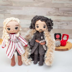 以下が含まれることがあります： 2体の編みぐるみ人形。1体は金髪で赤と白のストライプのドレスを着ており、もう1体は黒髪で灰色の毛皮の縁取りのあるマントを着ています。