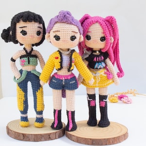 May include: Three handmade crochet dolls with different hairstyles and outfits. The dolls have black, purple, and pink hair, and wear colorful clothing, including jackets, shorts, and skirts. Each doll stands on a small wooden disc.