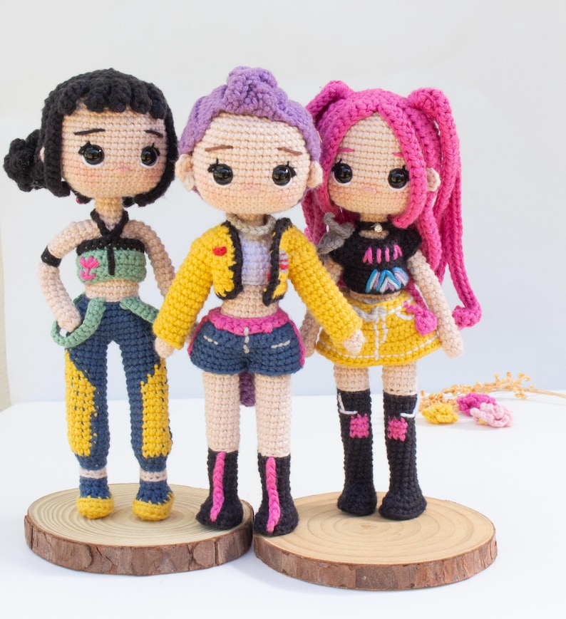 May include: Three handmade crochet dolls with different hairstyles and outfits. The dolls have black, purple, and pink hair, and wear colorful clothing, including jackets, shorts, and skirts. Each doll stands on a small wooden disc.
