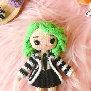 May include: A crocheted doll with green hair, a black and white striped jacket, a black skirt, and black and white striped socks. The doll is sitting on a pink fuzzy surface.