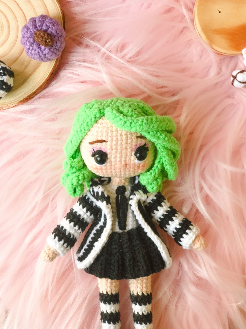 May include: A crocheted doll with green hair, a black and white striped jacket, a black skirt, and black and white striped socks. The doll is sitting on a pink fuzzy surface.
