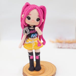 May include: A handmade crochet doll with pink hair styled in two long braids. The doll wears a black top, yellow skirt, and black boots. It stands on a small wooden base, showcasing a detailed and colorful design.