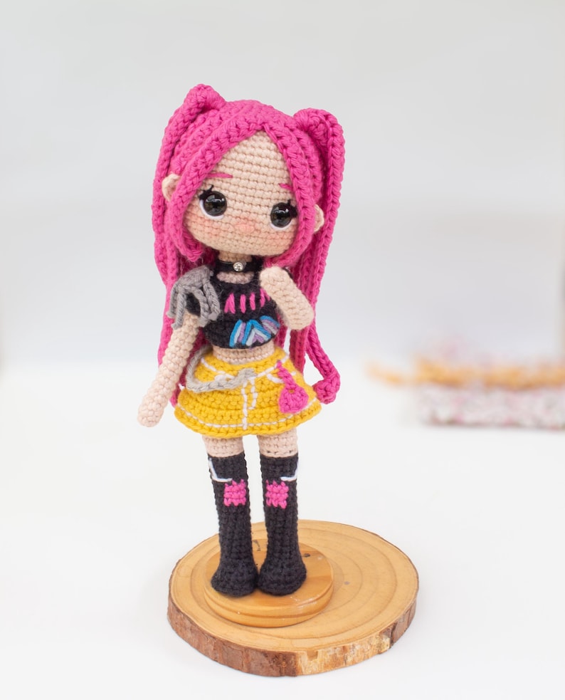 May include: A handmade crochet doll with pink hair styled in two long braids. The doll wears a black top, yellow skirt, and black boots. It stands on a small wooden base, showcasing a detailed and colorful design.
