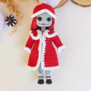 Puede incluir: Muñeca de ganchillo hecha a mano con un abrigo rojo con ribete blanco y un sombrero a juego. La muñeca tiene una cara gris, pelo rojo y ojos negros. El atuendo de la muñeca incluye pantalones grises y azules y zapatos negros.