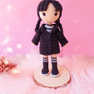 May include: Crocheted doll with black hair in braids, wearing a black coat over a white and black striped shirt, black boots with white trim, and white and black striped socks. The doll is displayed on a wooden slice.