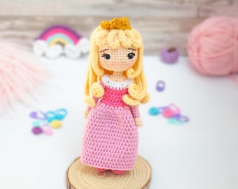 Könnte beinhalten: Eine gehäkelte Puppe mit langen blonden Haaren, die ein rosa Kleid und eine goldene Krone trägt. Die Puppe steht auf einem Holzsockel.