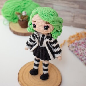 May include: A crocheted doll with green hair, wearing a black and white striped dress and jacket, and black and white striped stockings. The doll is standing on a wooden base.