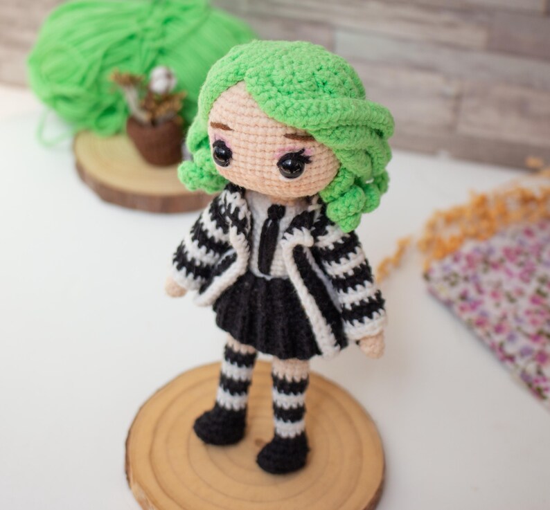 May include: A crocheted doll with green hair, wearing a black and white striped dress and jacket, and black and white striped stockings. The doll is standing on a wooden base.