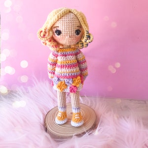 May include: A handmade crochet doll with blonde curly hair, rosy cheeks, and large black eyes. The doll wears a striped sweater in pink, yellow, and orange, a skirt with flower accents, and orange shoes. It stands on a wooden slice against a pink background.