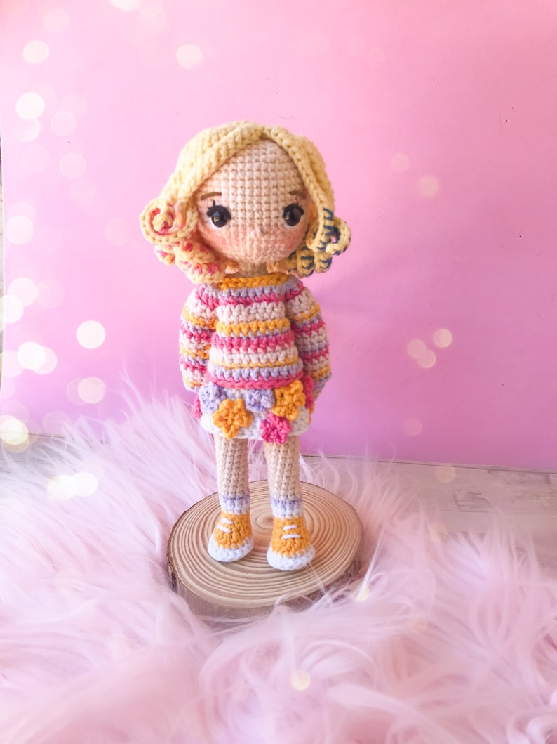 May include: A handmade crochet doll with blonde curly hair, rosy cheeks, and large black eyes. The doll wears a striped sweater in pink, yellow, and orange, a skirt with flower accents, and orange shoes. It stands on a wooden slice against a pink background.