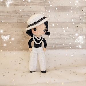 以下が含まれることがあります： 白い帽子に黒いリボン、黒いセーター、白い真珠のネックレス、白いズボンを着用した、かぎ針編みのドール。ドールは金色のキラキラが散りばめられた白い表面の上に立っている。