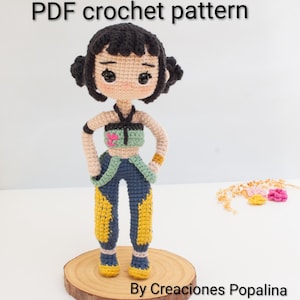 Puede incluir: Muñeca de ganchillo con cabello negro recogido en dos moños, vestida con un top verde y rosa y pantalones azules con detalles amarillos. La muñeca se exhibe sobre una base de madera. El texto "PDF crochet pattern" y "By Creaciones Popalina" está incluido.