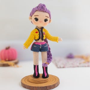 May include: A handmade crochet doll with purple hair, wearing a yellow jacket, denim shorts, and black boots with pink accents. The doll is standing on a wooden base, showcasing a unique, handcrafted design.
