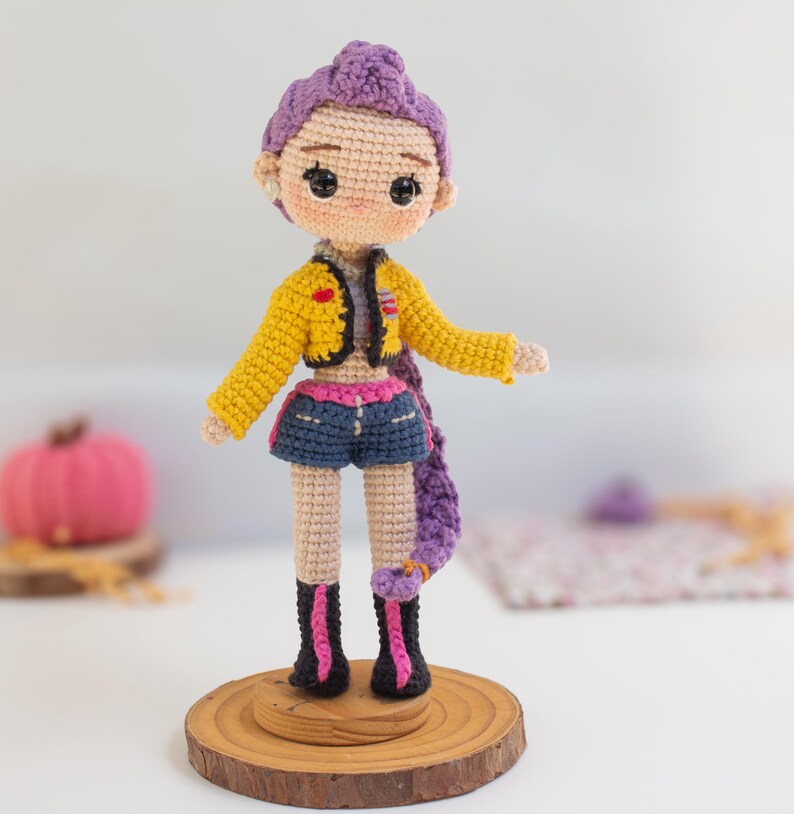 May include: A handmade crochet doll with purple hair, wearing a yellow jacket, denim shorts, and black boots with pink accents. The doll is standing on a wooden base, showcasing a unique, handcrafted design.