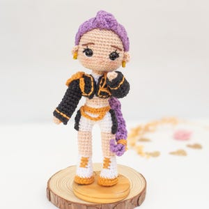 Op de afbeelding: Handgemaakte gehaakte pop met een paars kapsel, gouden oorbellen en een zwart-gouden outfit. De pop staat op een houten voet. De outfit omvat een zwart jasje met gouden rand, witte shorts en oranje en witte laarzen.