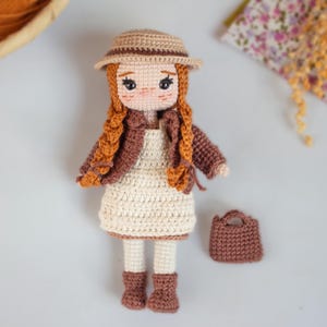 Puede incluir: Muñeca de ganchillo hecha a mano con cabello rojo trenzado, vestida con un vestido beige, chaqueta marrón y sombrero. La muñeca tiene un bolso y botas marrones a juego. La muñeca mide aproximadamente 20 cm de altura.