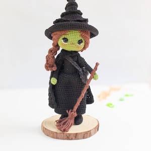 Puede incluir: Muñeca de bruja hecha a mano con ganchillo, con vestido, sombrero y capa negros. La muñeca tiene piel verde, pelo castaño trenzado y una escoba marrón. Se apoya en una base de rodaja de madera, ideal para Halloween.