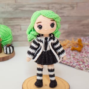 May include: A crocheted doll with green hair, a black and white striped jacket, a black skirt, and black and white striped stockings. The doll is standing on a wooden base.