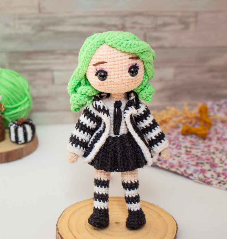 May include: A crocheted doll with green hair, a black and white striped jacket, a black skirt, and black and white striped stockings. The doll is standing on a wooden base.